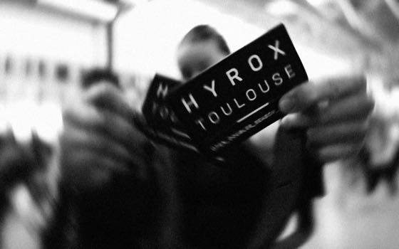 Carton d'une compétition Hyrox tenu à bout de bras, en noir et blanc